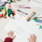 A child engages in creative drawing with colored pencils surrounded by wooden toys on a table.